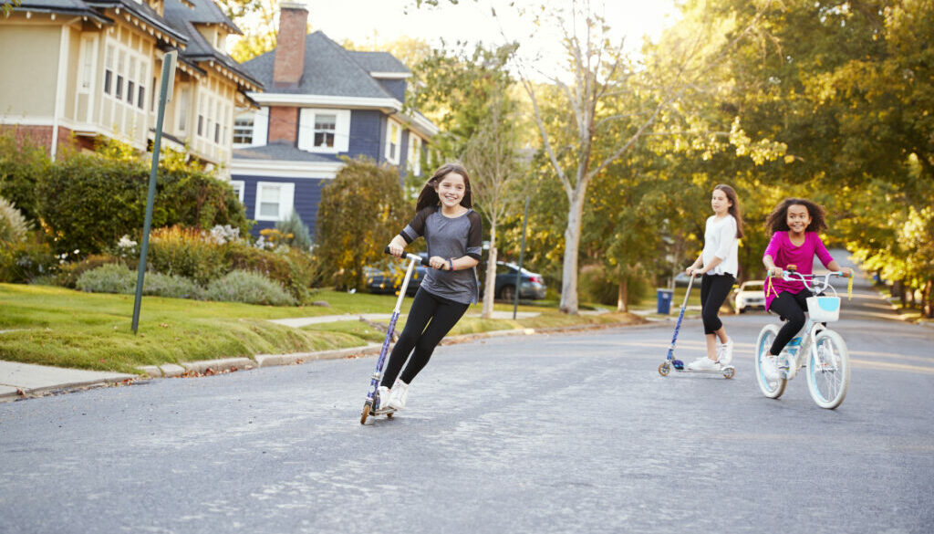 Three pre-teen girls playing in street on scooters and bike