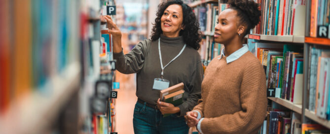 Mid adult Hispanic librarian escorting a student in a library. Searching for a book on shelve.