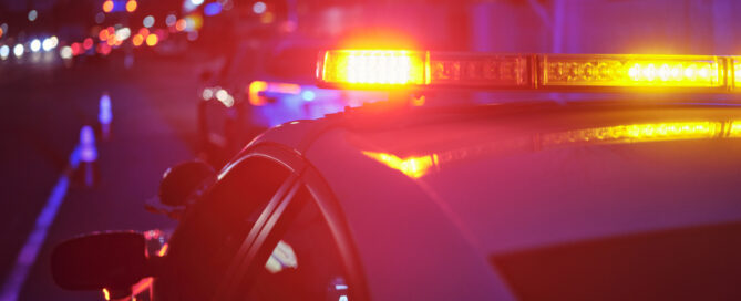 A police car at night alongside a road with emergency lights flashing.